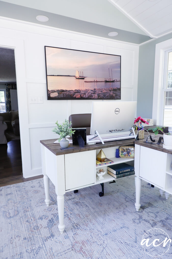 A bright, modern home office reveal featuring a large monitor on a desk, white paneled walls, and a wall-mounted photo of sailboats. Natural light streams in through the window, giving this sunroom-inspired space an airy and organized feel.