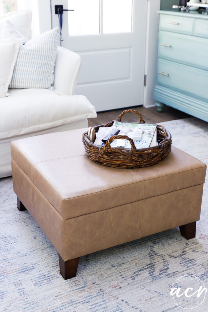 A tan leather ottoman sits on a patterned rug, topped with a woven tray holding magazines. Behind it, in this sunroom, there is a white couch with blue and white pillows, a blue dresser, and a door with sunlight streaming through the window.