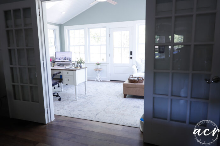 Bright home office sunroom with white French doors, a desk with a computer, a patterned rug, and a cozy sofa under large windows letting in natural light. A small round side table sits by the window.