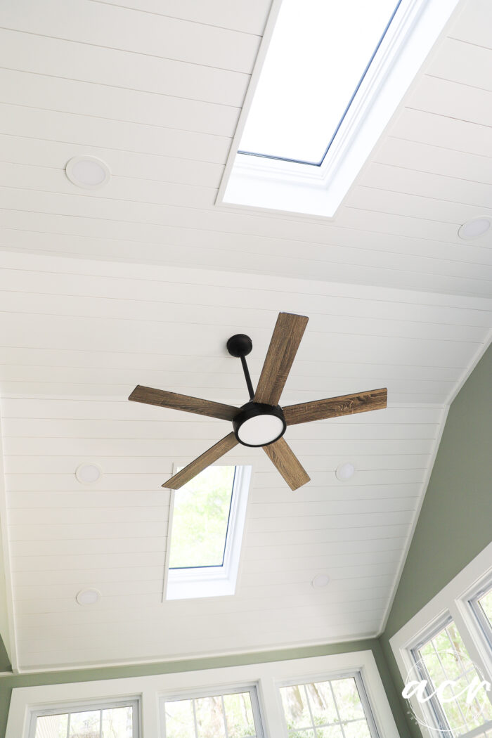 A white shiplap ceiling with two rectangular skylights and a modern ceiling fan with wooden blades brightens this sunroom, featuring green walls and large windows.