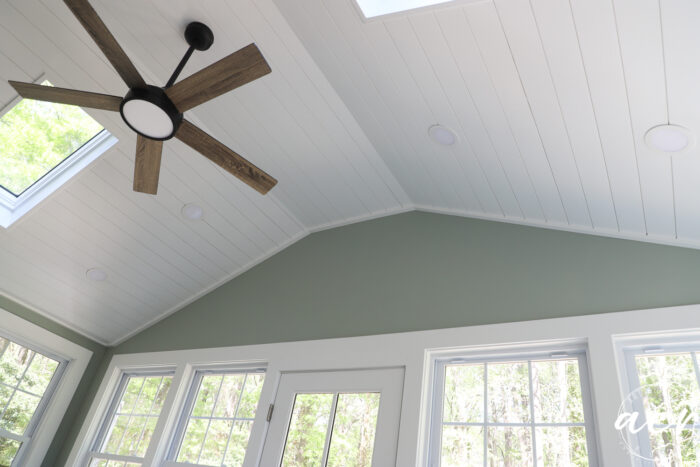 Sunroom Office Reveal: White shiplap ceiling with a modern ceiling fan, skylights, and recessed lights. Green accent wall with large windows and a door letting in natural light, showing trees outside.