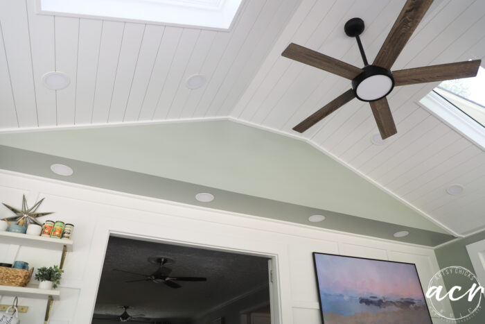 A white shiplap vaulted ceiling with a modern wood ceiling fan, skylights, and recessed lights brightens this sunroom. Below, a light green accent wall features shelves with decor and a landscape painting.