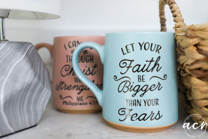 Two ceramic mugs sit on a countertop in a cozy sunroom. The front mug is light blue with the words Let your faith be bigger than your fears, while the pink mug features an inspirational Bible verse. A lamp and woven basket are nearby.