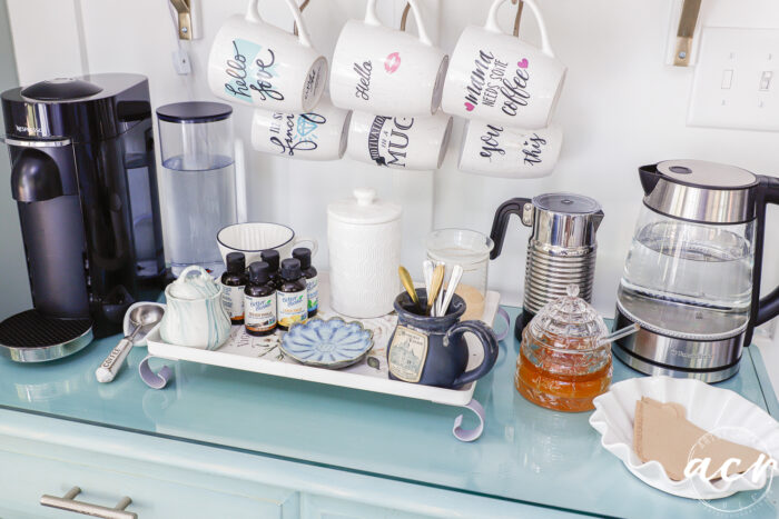 A cozy coffee station in the sunroom features mugs hanging above, a coffee maker, electric kettle, glass honey jar, coffee syrups, sugar jar, plates, and utensils arranged on a blue countertop.