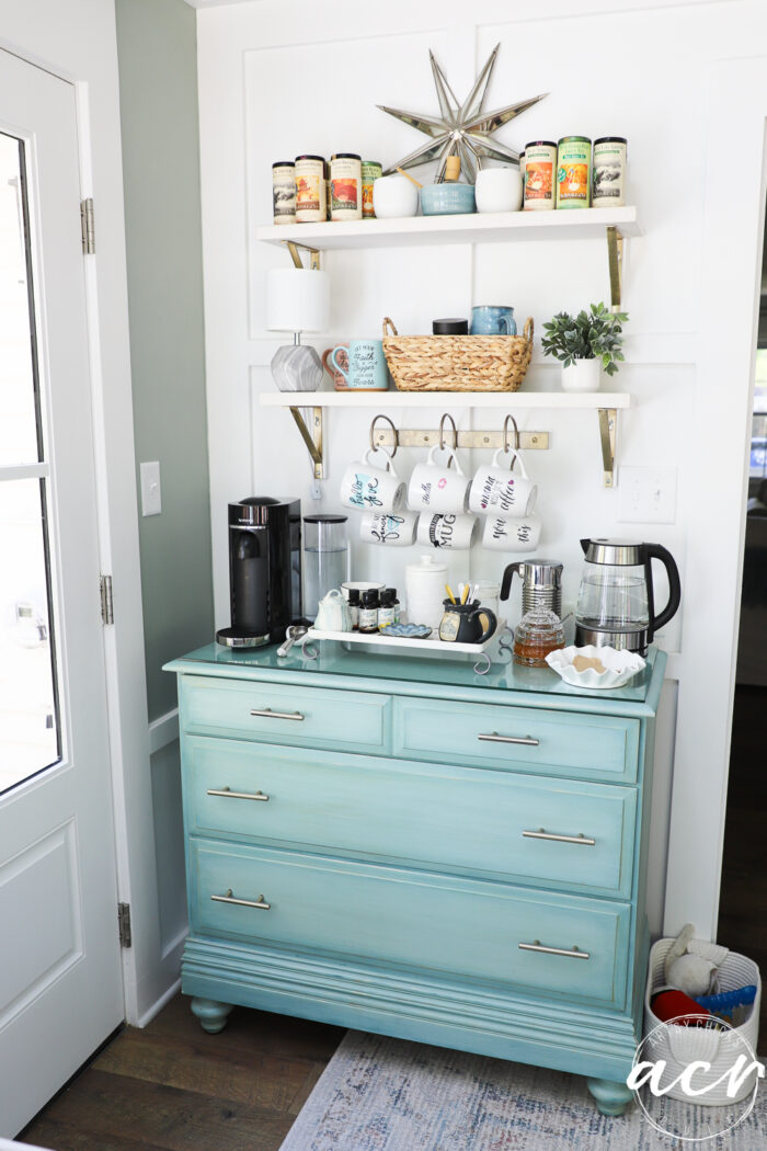 A light blue dresser in the sunroom holds a coffee station with a coffee maker, mugs, glasses, and supplies. Above, two white shelves display cans, mugs, a basket, and a plant. A starburst decor hangs on the white wall.
