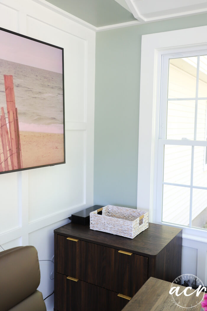 A corner of a modern sunroom with a window, wooden dresser with gold handles, a woven basket on top, a wall-mounted beach photo, and light green and white walls.