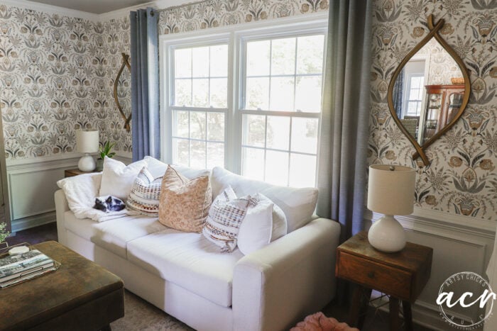 A bright living room with a white couch, patterned pillows, and a small dog lying on the armrest. Unique mirrors adorn the walls, complementing the floral wallpaper, wooden side table, and gray-curtained windows.
