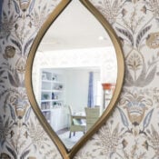 Gold-framed oval mirror with organic curved ends, mounted on floral-patterned wallpaper; its reflection reveals a light-filled room with a green chair, shelving, and blue curtains.