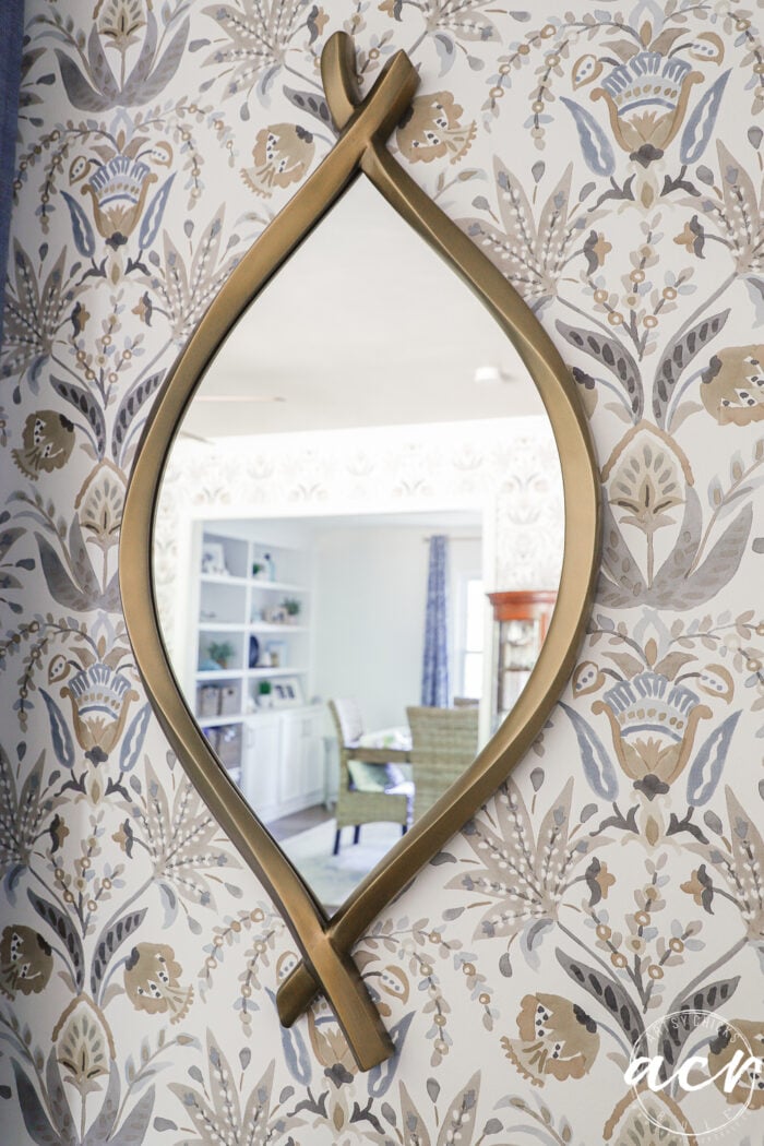Gold-framed oval mirror with organic curved ends, mounted on floral-patterned wallpaper; its reflection reveals a light-filled room with a green chair, shelving, and blue curtains.