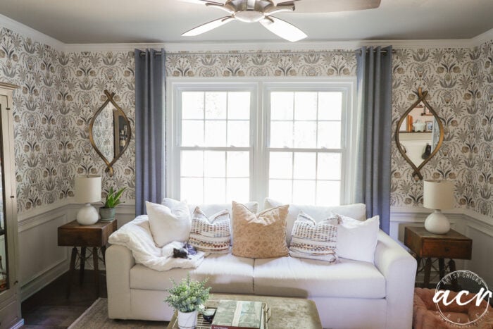 Bright living room with a white couch, neutral throw pillows, side tables with lamps, organic curved mirrors on patterned wallpaper, a ceiling fan, and large windows with gray curtains letting in natural light.
