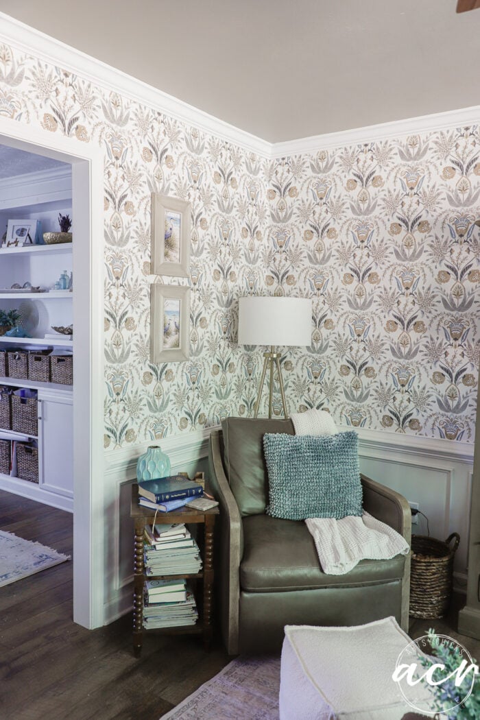 A cozy reading nook with a gray armchair, blue pillows, a side table stacked with books, a modern floor lamp, and floral wallpaper. Organic curved mirrors add flair above the white shelving with baskets and decor in the adjacent room.