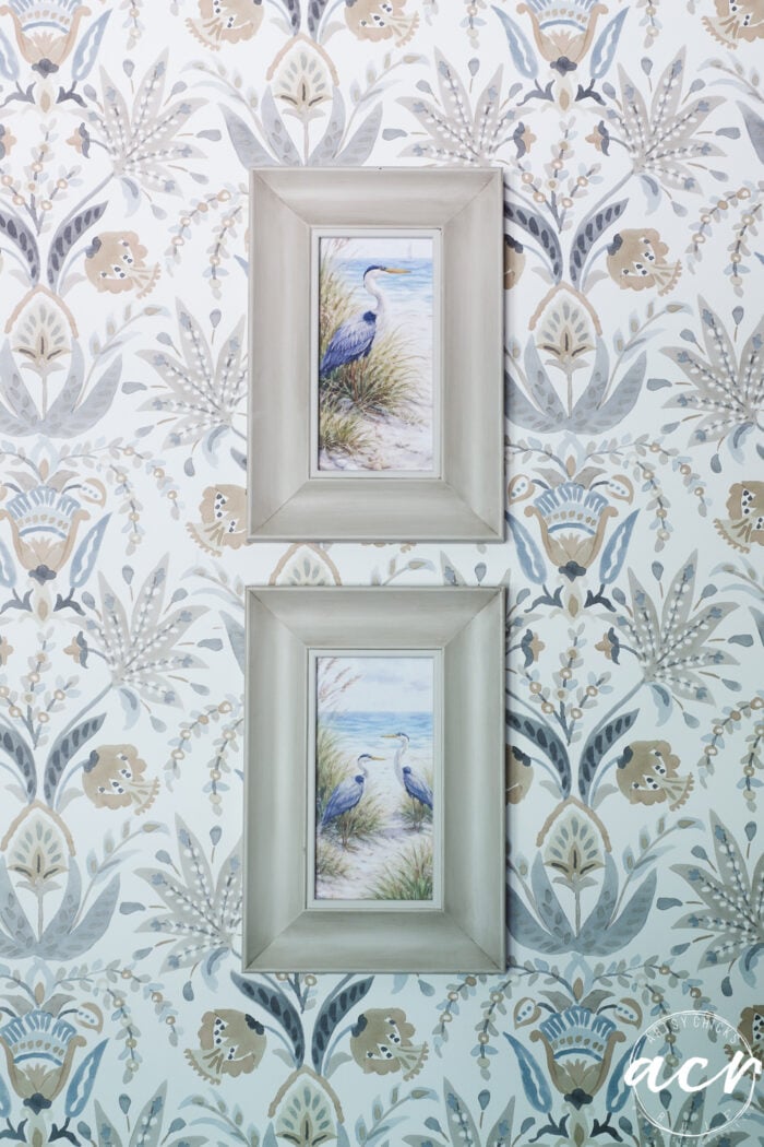 Two framed paintings of blue herons, one above the other, hang on a wall with floral-patterned wallpaper in soft pastel shades of blue, beige, and gray, next to organic curved mirrors that add a modern touch to the serene space.