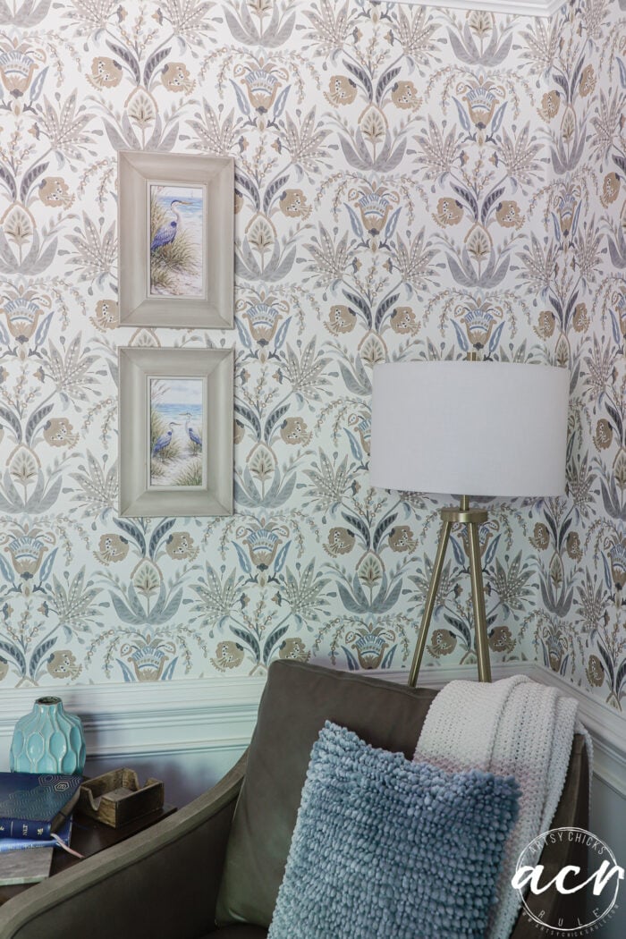A cozy corner with a gray armchair, a textured blue throw pillow, organic curved mirrors reflecting light, a tripod floor lamp, two framed beach-themed pictures on floral patterned wallpaper, and small items on a side table.