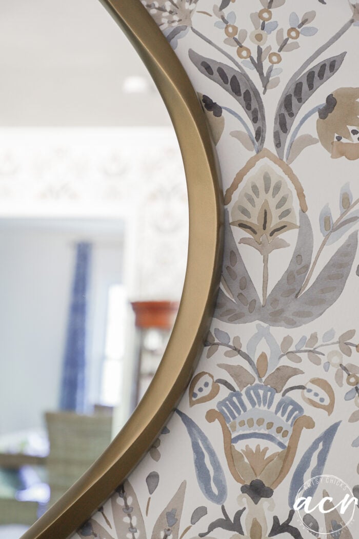 A close-up of an organic curved mirror with a gold frame, reflecting a room with blurred furniture. The wall behind the mirror features decorative floral and leaf patterned wallpaper in muted blue, gray, and beige tones.