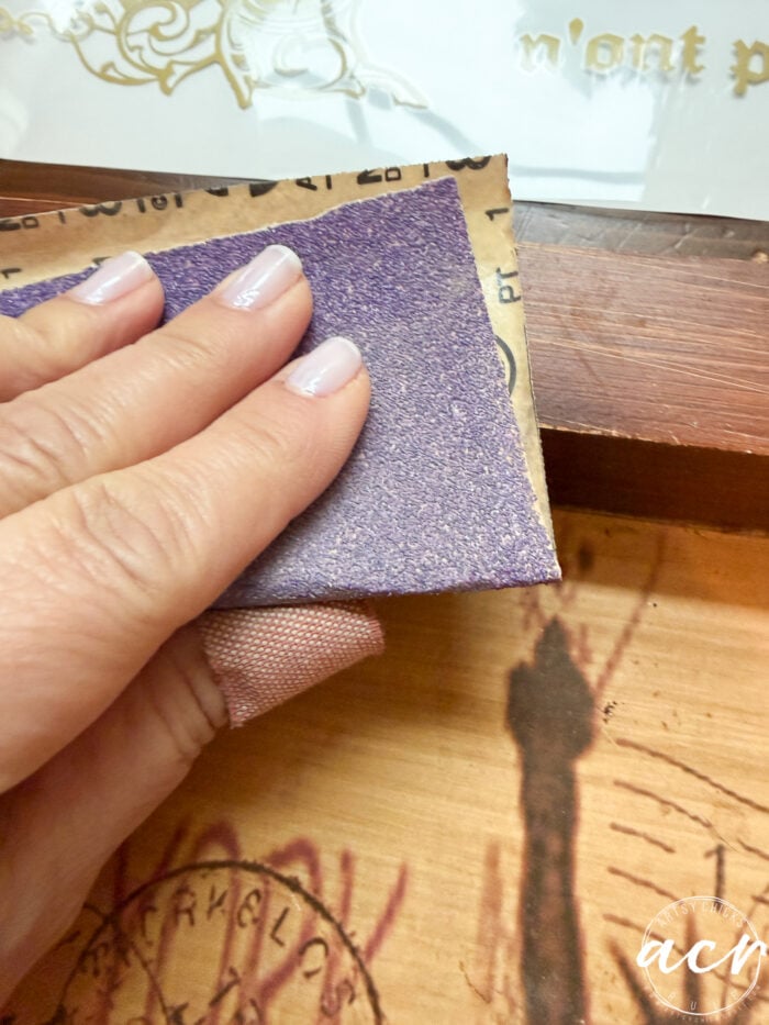A hand with light-colored nail polish holds a piece of sandpaper, carefully sanding the edge of a wooden tray with a stamped or printed design&mdash;perhaps a unique find from a Thrift Shop.