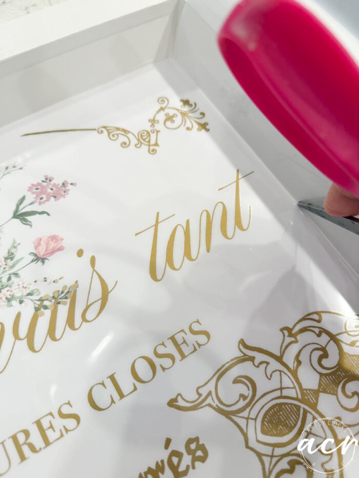 Close-up of a person applying gold vinyl lettering with a scraper tool onto thrift shop trays, featuring floral and ornate decorative designs.