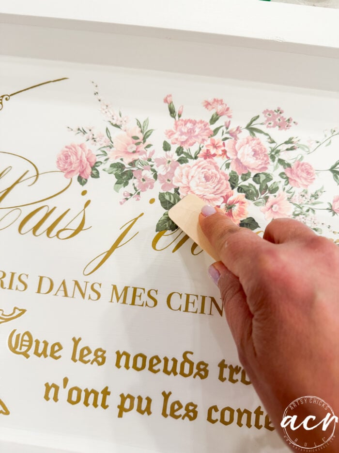 A hand uses a tool to apply a floral decal with pink flowers and green leaves onto a white tray from a thrift shop, accented by gold French text.