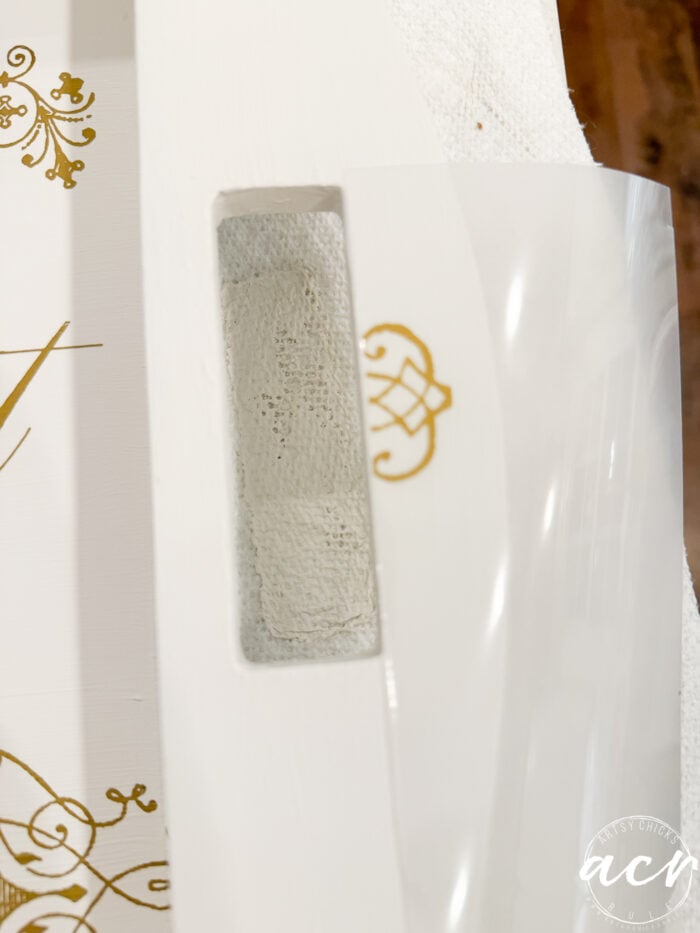 A close-up of a white wooden surface with a rectangular cut-out handle, partially covered by a clear plastic sheet with gold decorative designs from Two Trays, and a monogram visible on the plastic&mdash;perfect for a thrift shop find.