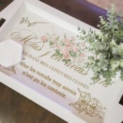 A white tray with floral designs and French text sits on a brown surface, as if found at a charming thrift shop. A small white hexagonal box and a green potted plant rest atop the tray, which features elegant gold script and pink flowers.