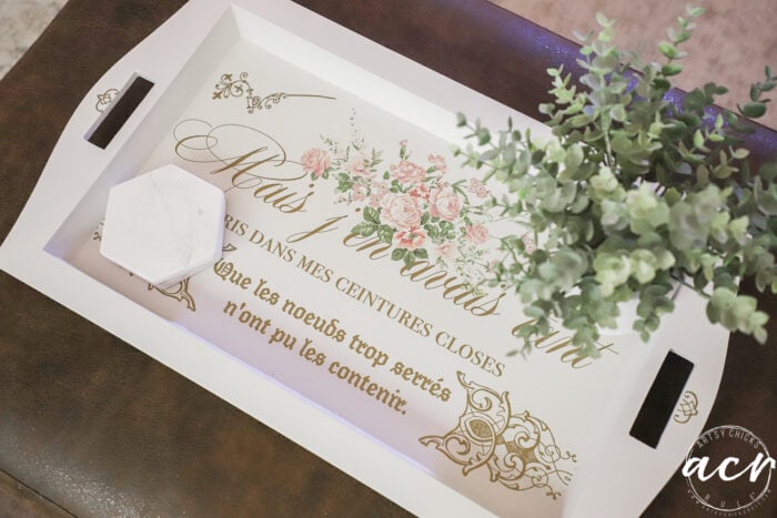 A white tray with floral designs and French text sits on a brown surface, as if found at a charming thrift shop. A small white hexagonal box and a green potted plant rest atop the tray, which features elegant gold script and pink flowers.