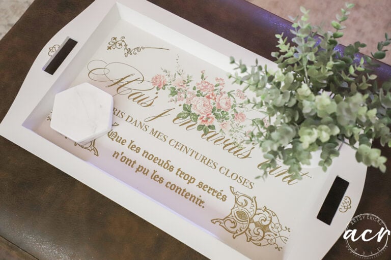 A white tray with floral designs and French text sits on a brown surface, as if found at a charming thrift shop. A small white hexagonal box and a green potted plant rest atop the tray, which features elegant gold script and pink flowers.