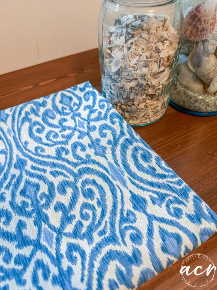 A blue and white patterned fabric or notebook from a thrift shop rests on a wooden surface next to two glass jars filled with seashells and small shells. The lower right corner features a white circular watermark.