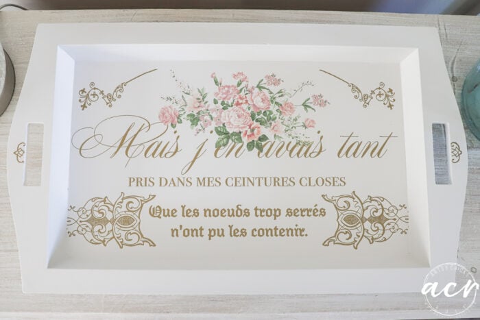 A white rectangular tray with side handles, decorated with pink flowers and gold French text in elegant script and print, sits on a light wooden surface. The lower right corner features a circular acy logo&mdash;perfect for chic trays from your favorite thrift shop.