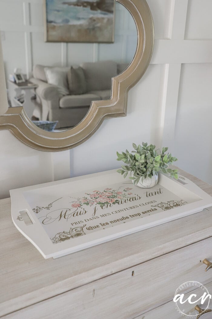 A decorative tray with floral designs and French text, found at a local thrift shop, rests on a light wooden dresser beside a small potted plant. A round mirror with a gold frame hangs above, reflecting a couch in the background.