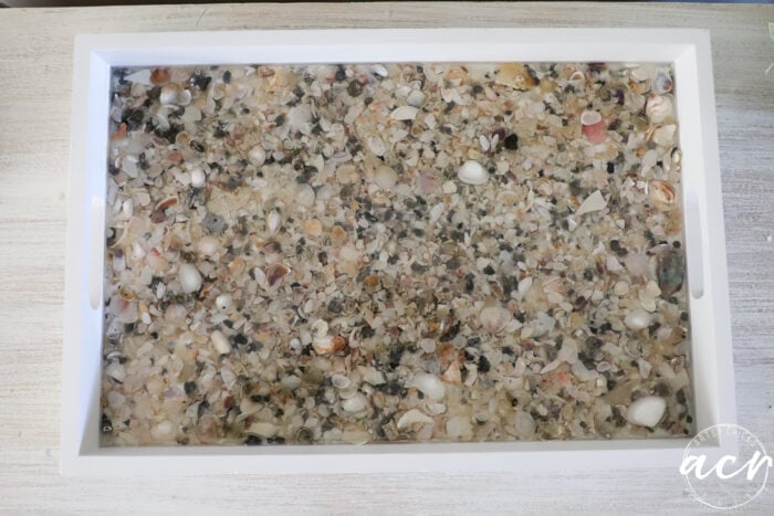 A white rectangular tray, perfect for trays lovers, filled with small, assorted seashells and pebbles embedded in clear resin, displayed on a light wood surface.