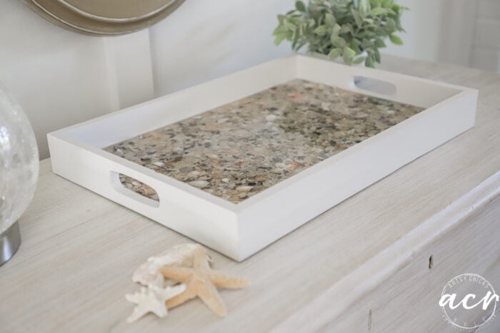 A rectangular white tray with handles sits on a light wooden surface. The tray&rsquo;s base is filled with small seashells and pebbles set in resin. Nearby, two trays from a thrift shop display more seashells, starfish, and a potted green plant.