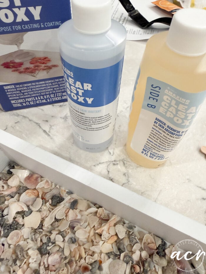 Two bottles labeled &ldquo;Clear Cast Epoxy&rdquo; sit on a marble countertop beside a thrift shop find&mdash;a seashell-covered tray. The bottles, marked Side A and Side B, rest near an epoxy box partially visible in the background.