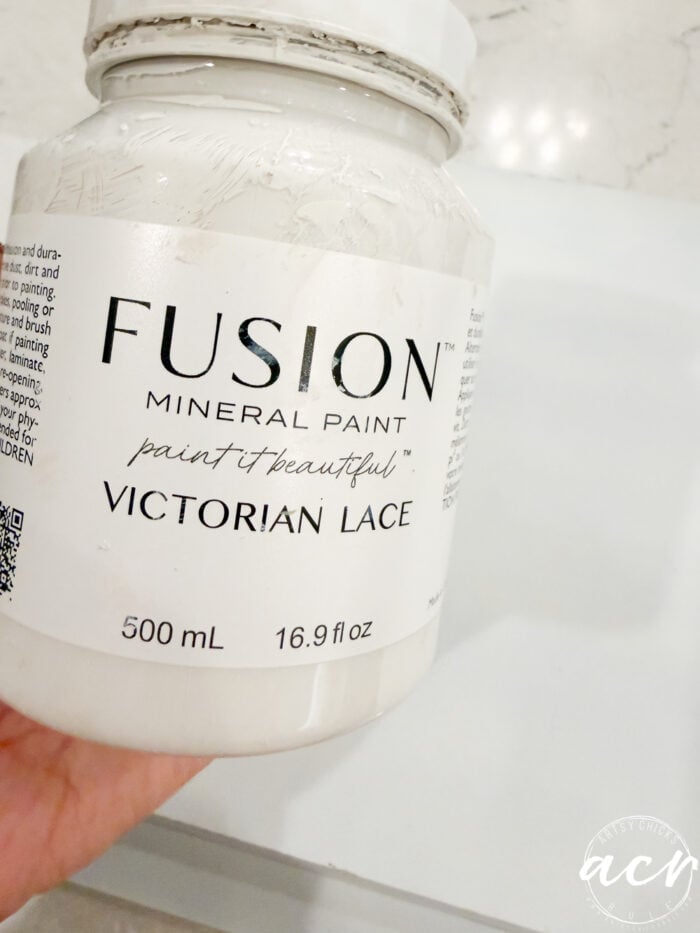 A hand holds a jar of Fusion Mineral Paint in Victorian Lace, perfect for upcycling thrifted trays or adding charm to your home decor. The label reads "paint it beautiful," 500 mL (16.9 fl oz), against a white background with a logo in the bottom right corner.