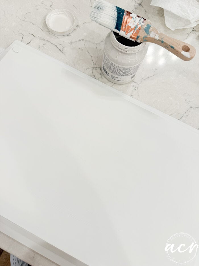 A paintbrush with white paint rests on an open paint jar, next to a freshly painted white surface on a marble countertop&mdash;perhaps supplies found at a Thrift Shop. A white lid and a piece of paper towel are also visible.