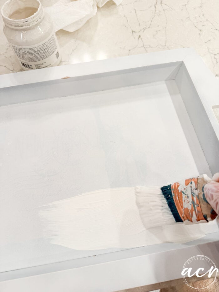 A hand holding a paintbrush applies white paint to the surface of a rectangular thrift shop tray on a light-colored countertop; an open paint jar sits nearby.