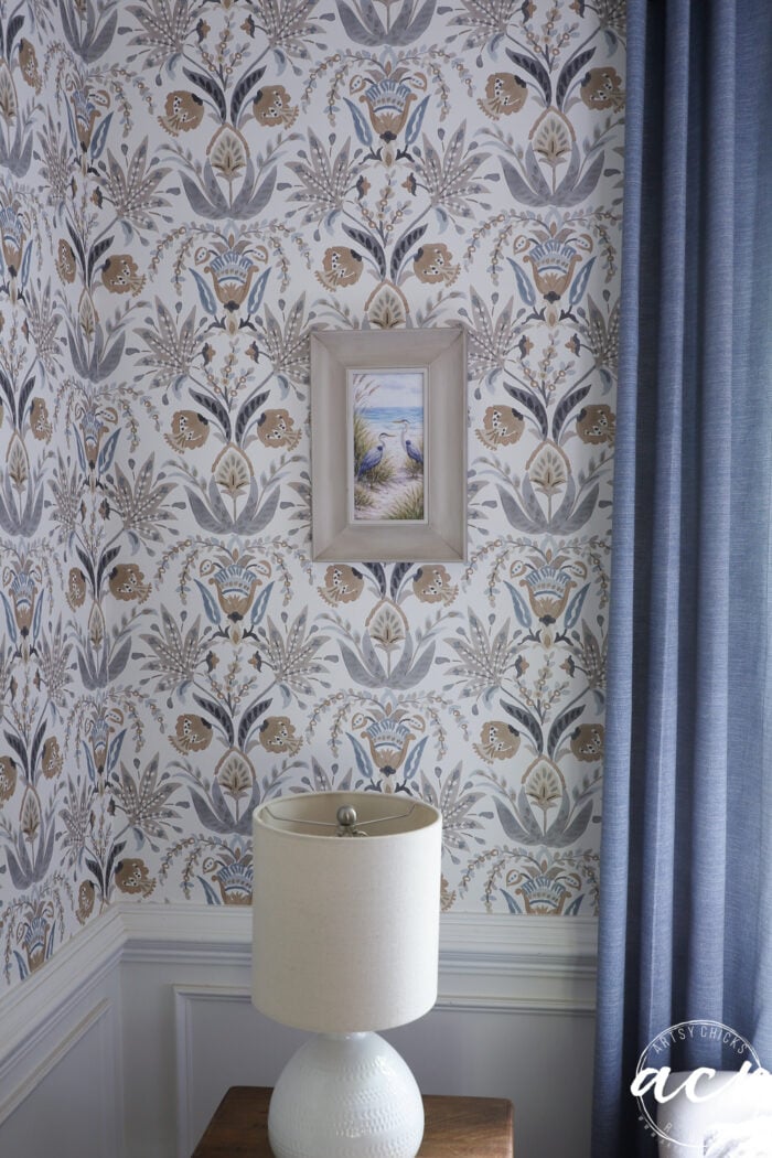 A corner of a room with floral-patterned wallpaper, a small upcycled framed print of a beach painting, a white textured lamp on a wooden table, gray wainscoting, and blue curtains on the right.