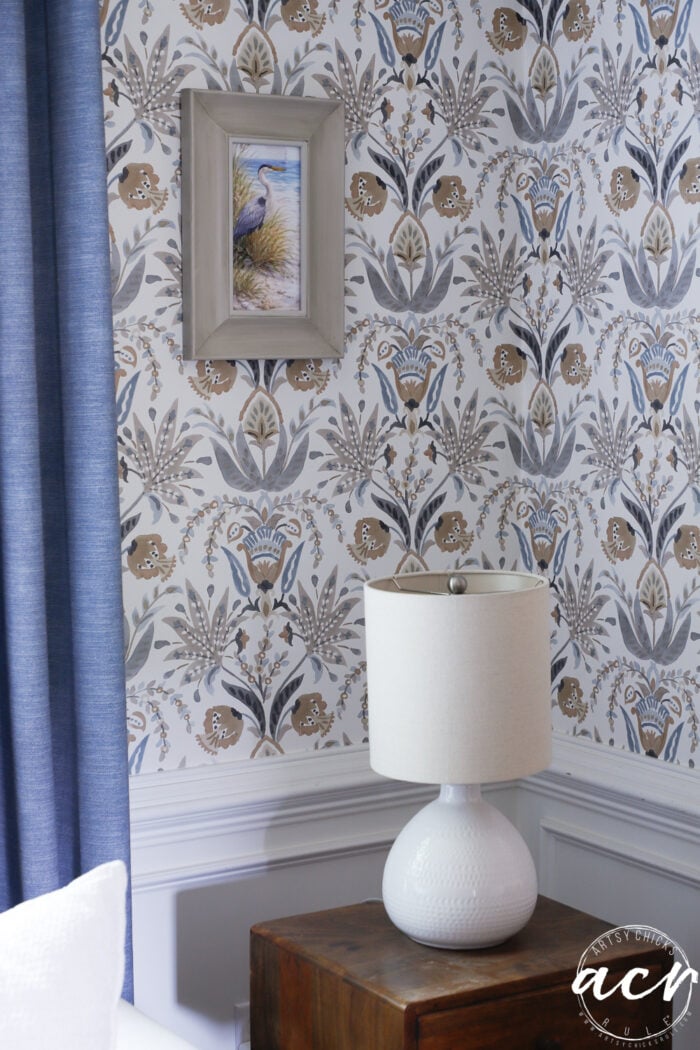 A white table lamp on a wooden side table sits in front of patterned wallpaper with blue, gray, and gold floral designs. Above the lamp is an upcycled framed print of a bird illustration, and a blue curtain hangs to the left.