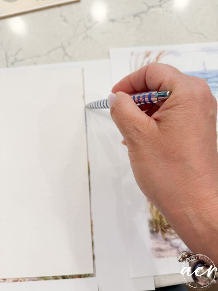 A hand holding a striped pen traces the edge of a paper atop another sheet featuring a beach scene Print Set, all arranged on a light-colored, marbled surface.