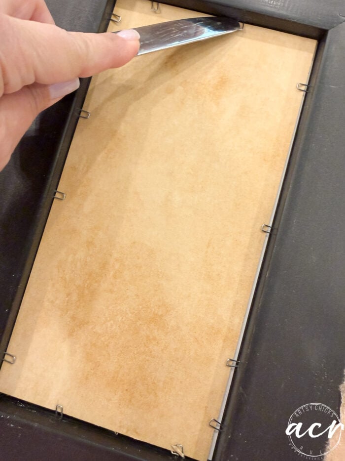 A hand uses a flat tool to lift metal tabs securing the back panel of a black framed print, preparing to remove the panel.