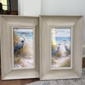 This print set features two framed prints of herons standing among beach grass and sand, with the ocean and sky behind. The light wood frames rest on a hardwood floor beside a wall, adding an elegant touch to any space.
