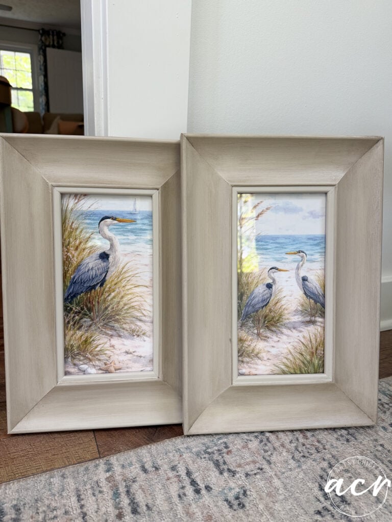 This print set features two framed prints of herons standing among beach grass and sand, with the ocean and sky behind. The light wood frames rest on a hardwood floor beside a wall, adding an elegant touch to any space.