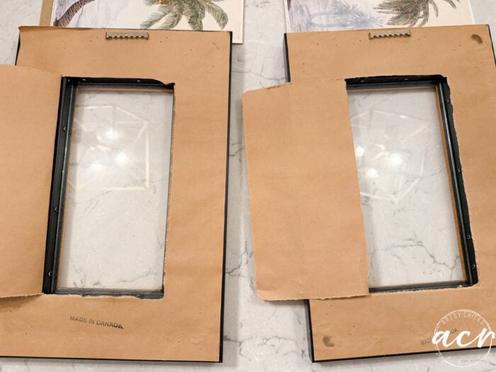 Two upcycled picture frames with torn brown paper backing lie face down on a marble surface. The frames have glass fronts, and peeling paper reveals the inside. The bottom left framed print says MADE IN CANADA.