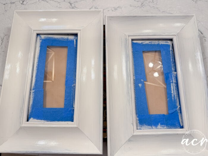 Two upcycled rectangular picture frames with glass centers covered by blue painter’s tape are shown side by side on a marble surface. The white frames appear to be freshly painted, with brush strokes visible.