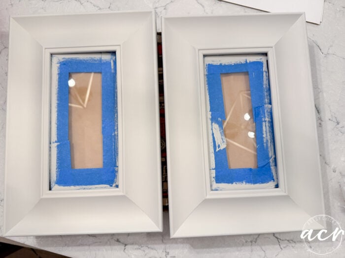 Two white picture frames on a marble surface, each with blue painters tape covering the glass center—perfect for an upcycled DIY project. Reflections of overhead lights are visible on the glass.