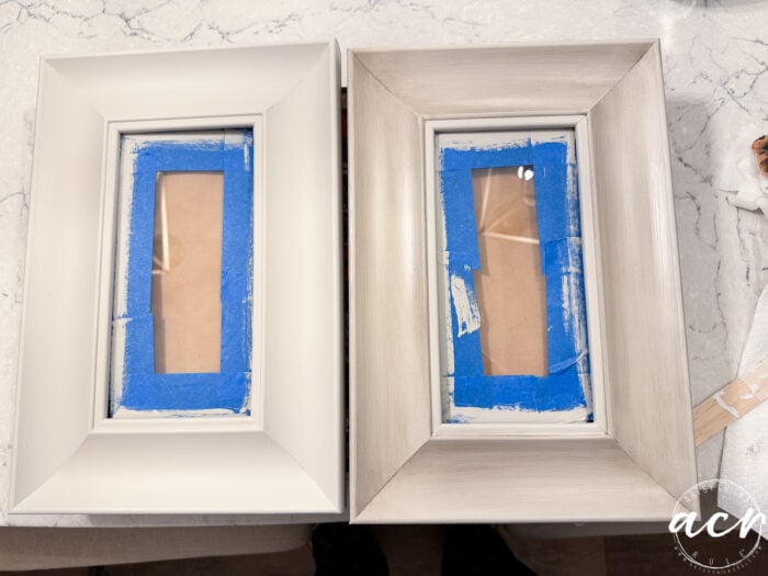 Two rectangular picture frames—an upcycled white frame and a weathered, grayish one—sit side by side on a white surface, blue painter’s tape covering the glass. Perfect for showcasing your favorite print set or framed print.