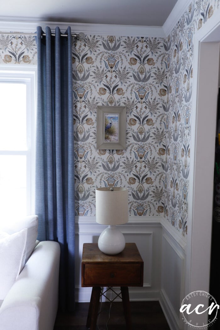 A cozy corner of a living room with patterned wallpaper, a framed print, a table lamp on an upcycled wooden side table, blue curtains, and part of a white couch.