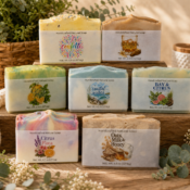 Seven handcrafted cold process soaps are displayed on a wooden surface with greenery, woven baskets, and bath accessories in the background. Each soap features a unique label and colorful design, highlighting the artistry of cold process soap making.