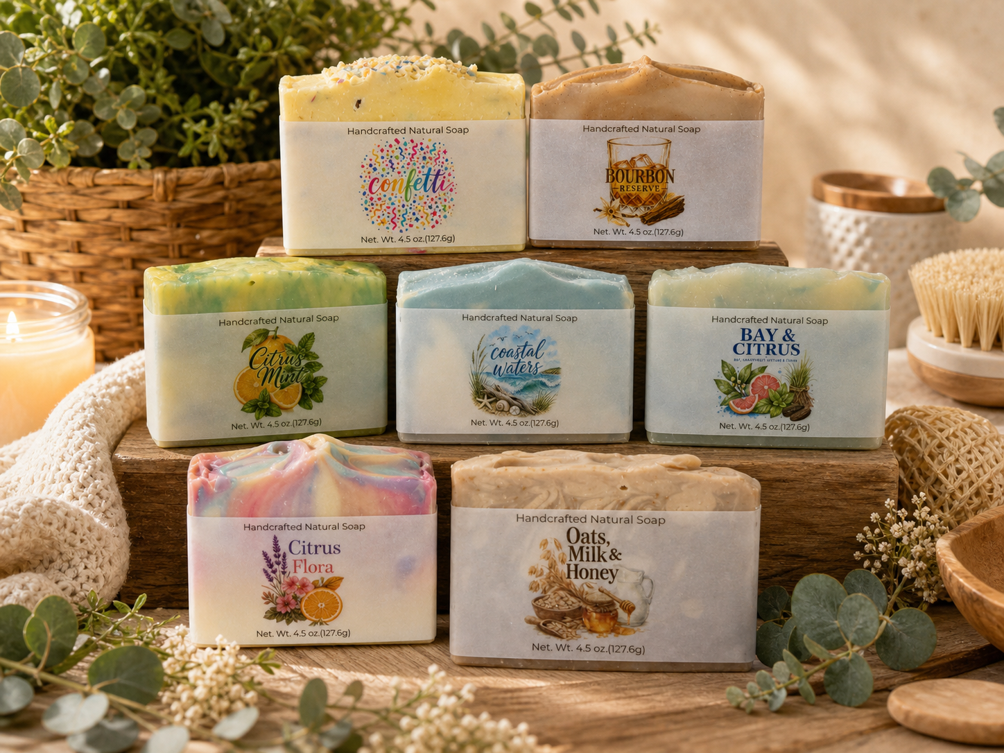 Seven handcrafted cold process soaps are displayed on a wooden surface with greenery, woven baskets, and bath accessories in the background. Each soap features a unique label and colorful design, highlighting the artistry of cold process soap making.