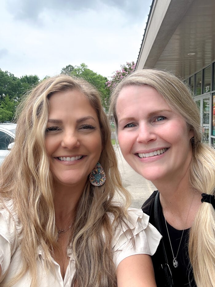 Two women smile for a selfie outside on a cloudy day, standing near a thrift shop with glass windows. One has wavy blonde hair and earrings; the other, in black, has straight blonde hair. Trees and parked cars are in the background.