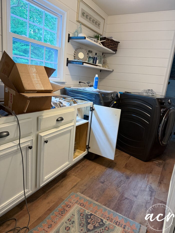 A recent Laundry Room Update features white cabinets, an open cabinet door, cardboard boxes on the counter, a black washer and dryer, open shelves with various items, and a window revealing greenery outside.