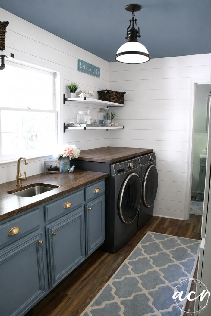 A stylish Laundry Room Update featuring blue cabinets, a wooden countertop, a sink, front-loading washer and dryer, open shelves with baskets, white shiplap walls, a blue ceiling, and a patterned rug on wood flooring.